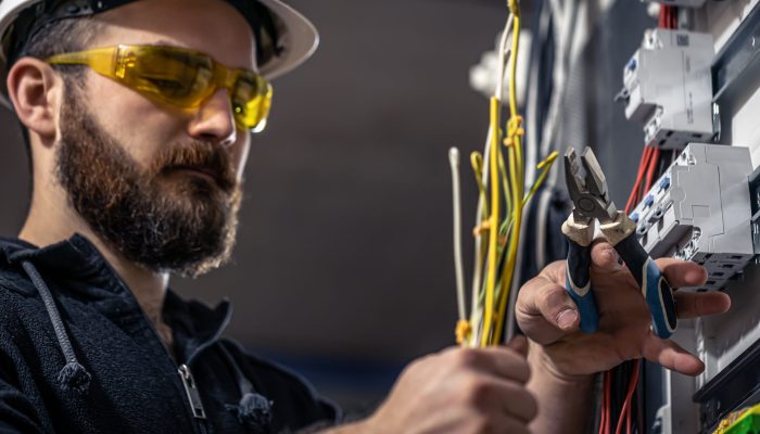 A male electrician works in a switchboard with an electrical connecting cable, connects the equipment with tools.